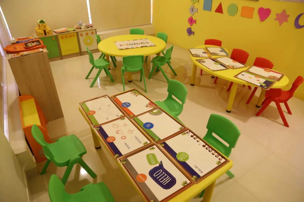 A preschool classroom with low yellow tables, green and red chairs, and worksheets laid out at each seat