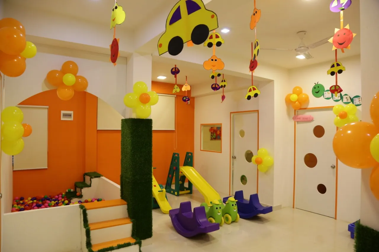 A decorated preschool play zone with orange walls, hanging balloon decorations, rockers, and a small slide