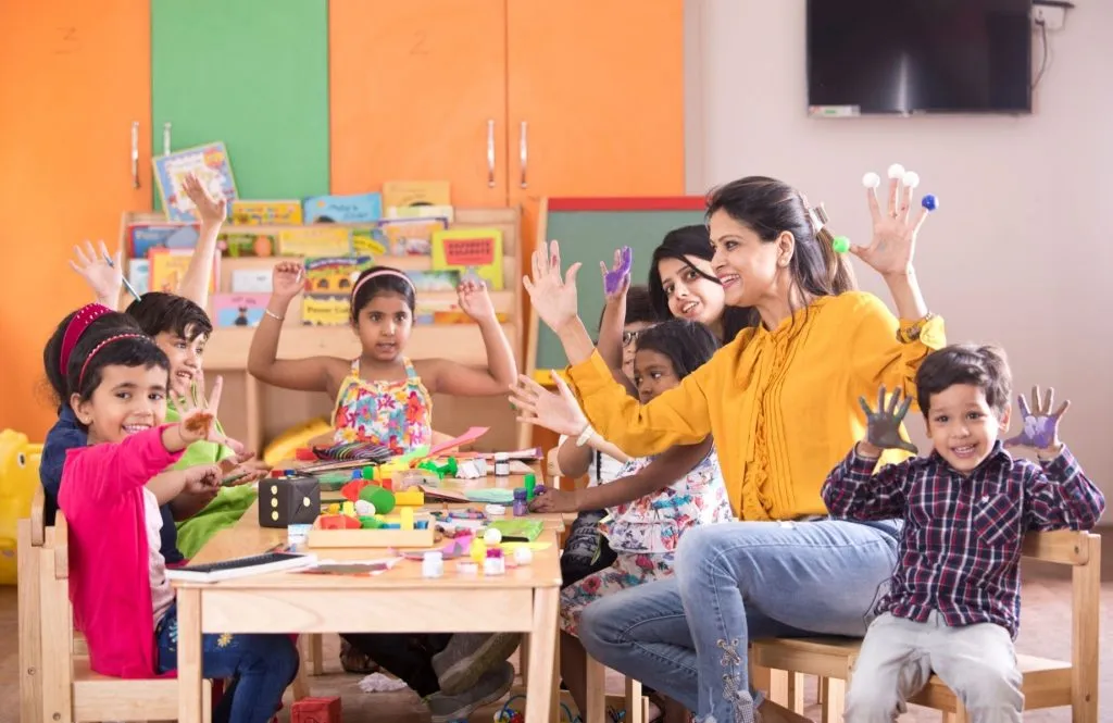 A preschool classroom with a low blue table, blue chairs, a whiteboard, and green storage shelves